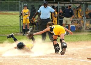 Girl softball catcher wearing protective gear tags runner at home plate, while the umpire , coach and teamates watch.